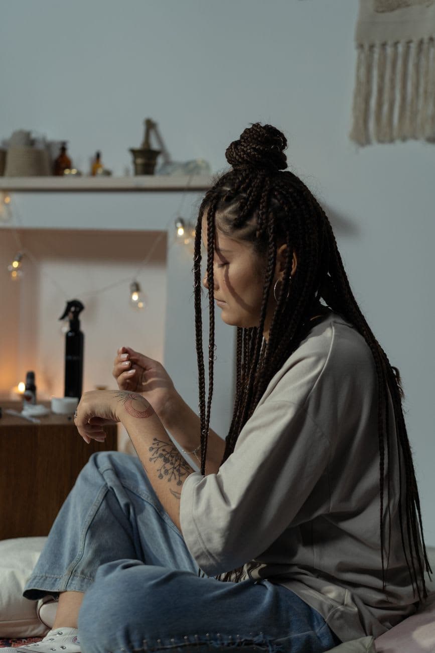 A woman sits indoors with braided hair, showcasing an arm tattoo in a serene setting.