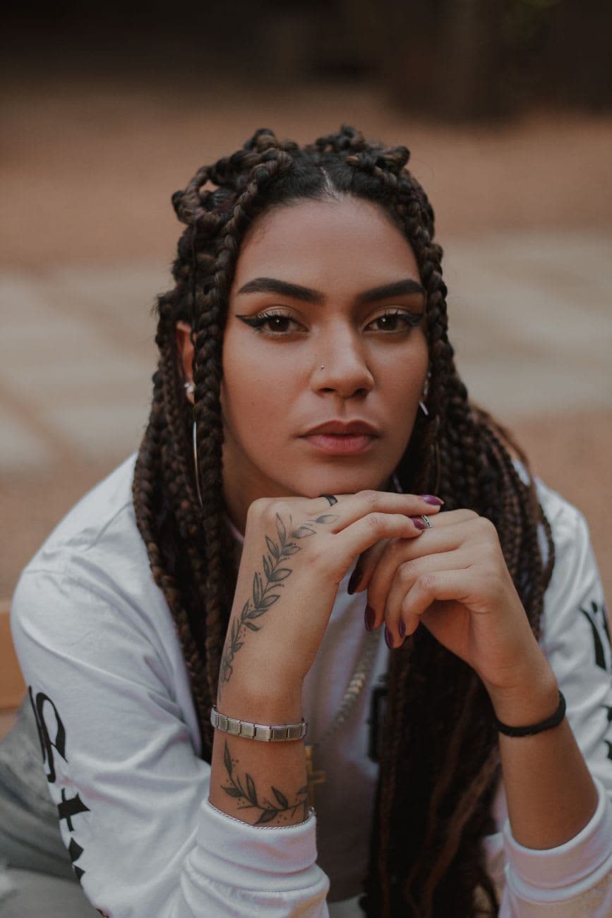A young woman with braided hair poses confidently outdoors, showcasing her tattoo and earrings.