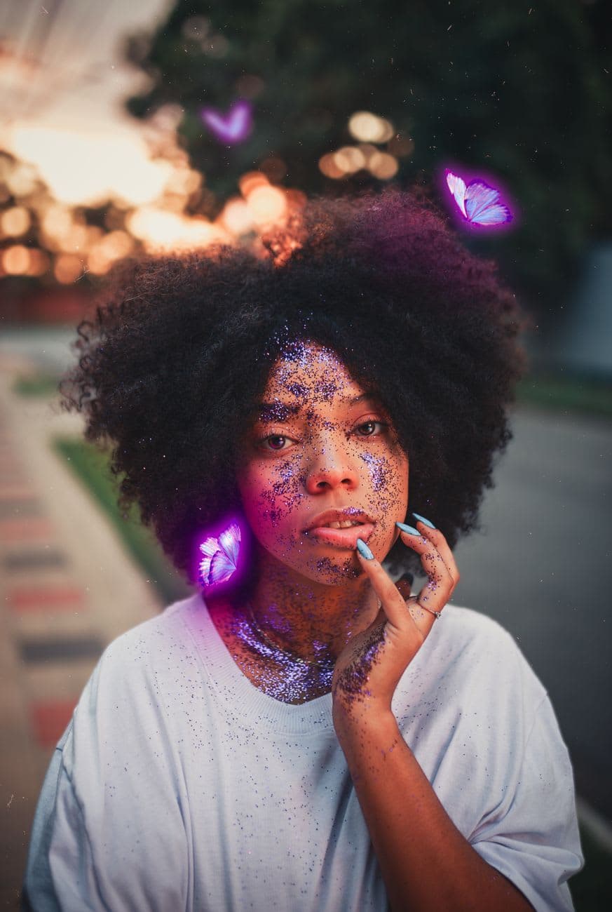 Artistic portrait of woman with afro hair, adorned with glitters and butterflies, at twilight.