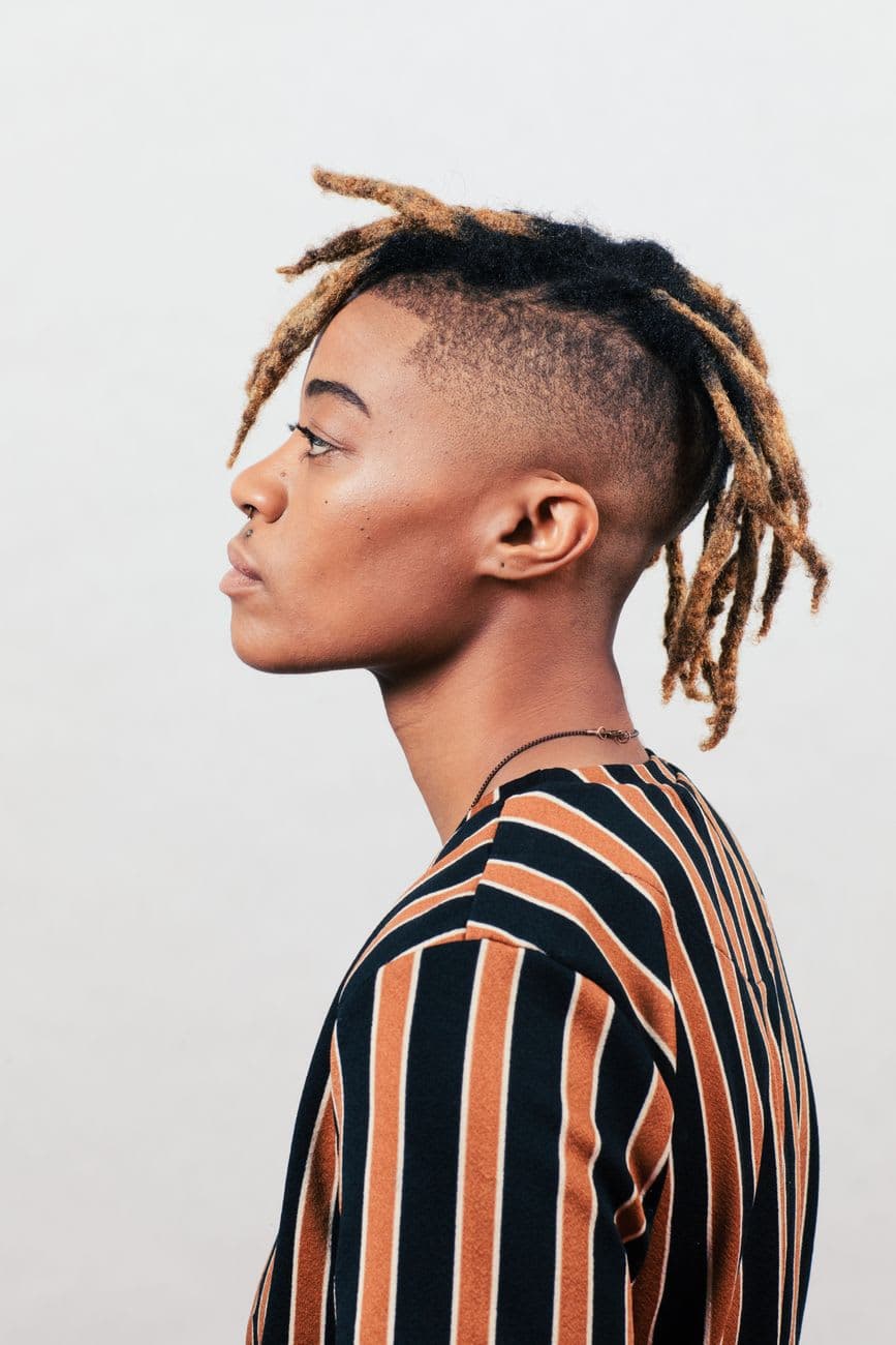 Profile portrait of a young person with distinctive loc hairstyle wearing a striped shirt against a white background.