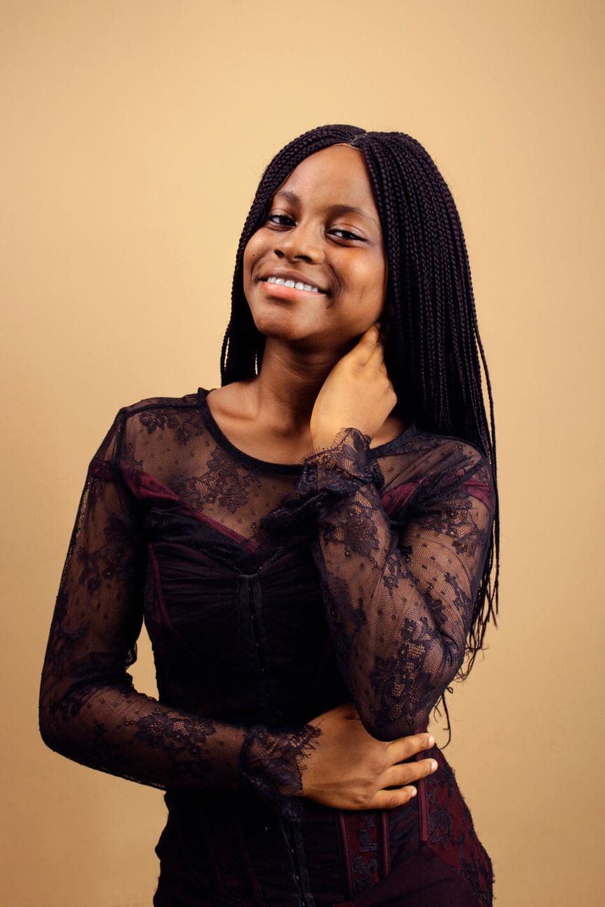 Stylish portrait of a young woman with long braids in a sheer black top against a brown background.