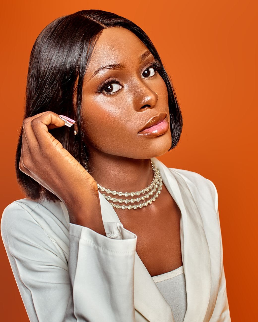 Portrait of a stylish black woman with pearl necklace and white blazer against an orange backdrop.