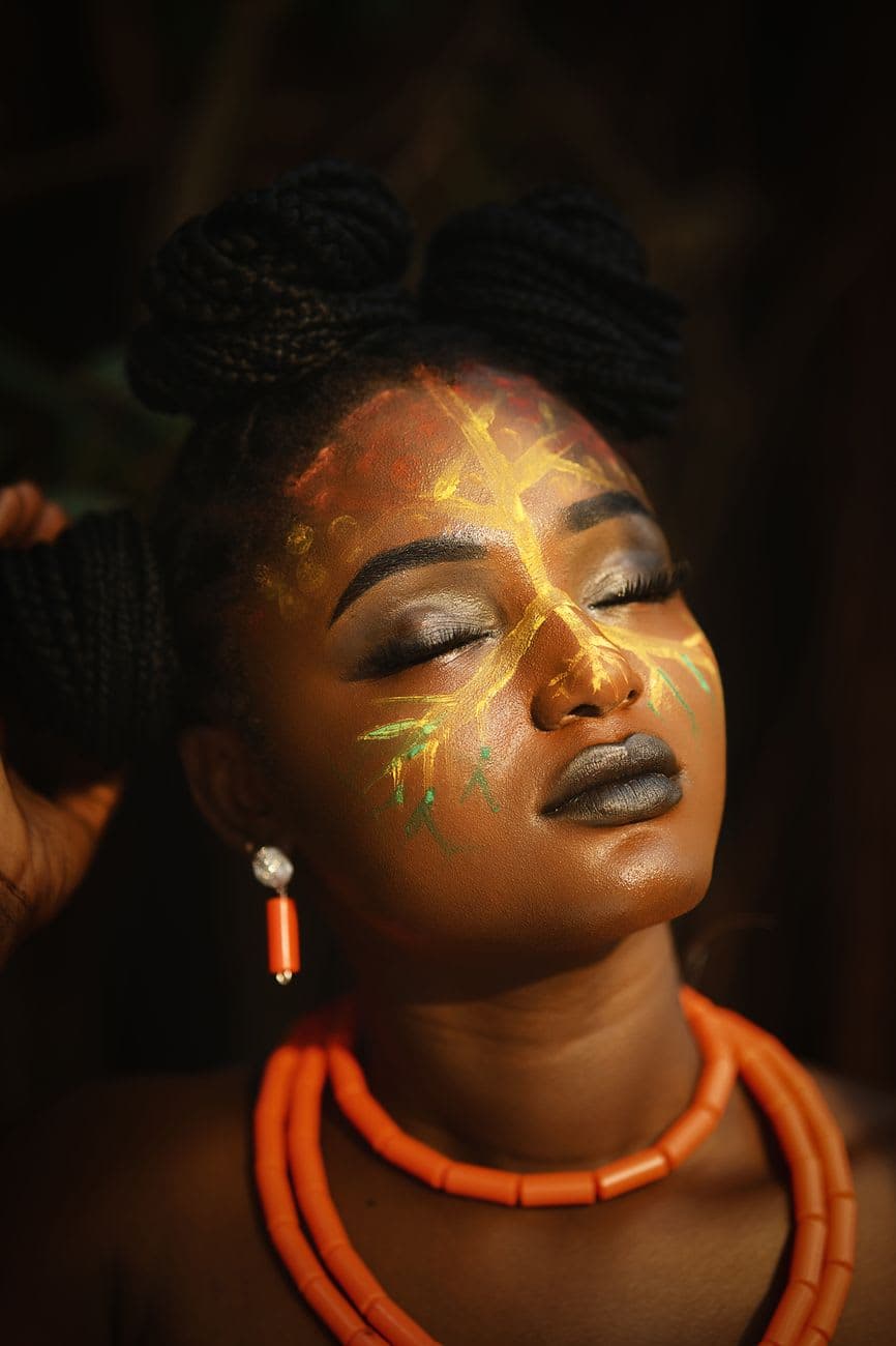African woman with vibrant face paint and beads, eyes closed, showing cultural expression.