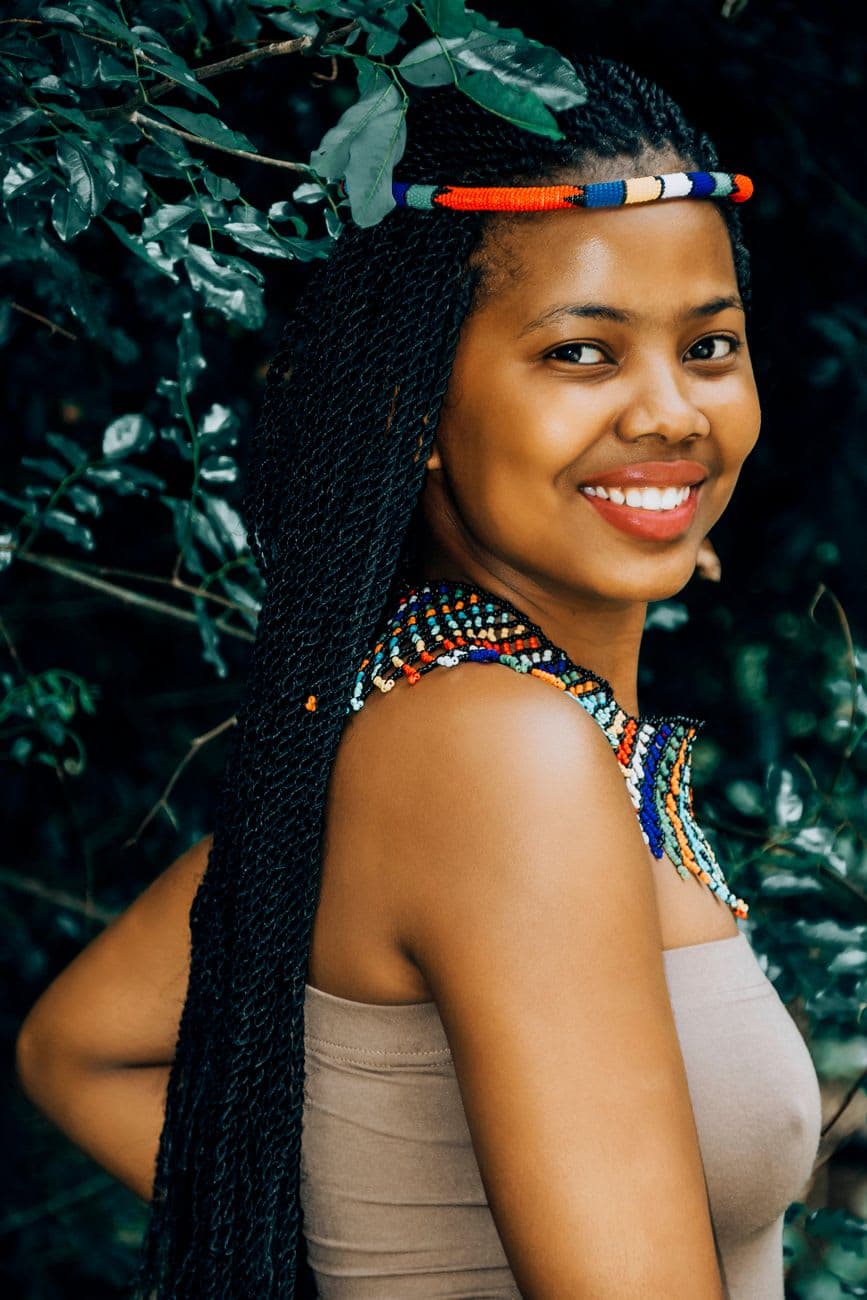 Smiling woman with long braids and colorful bead accessories in an outdoor setting.