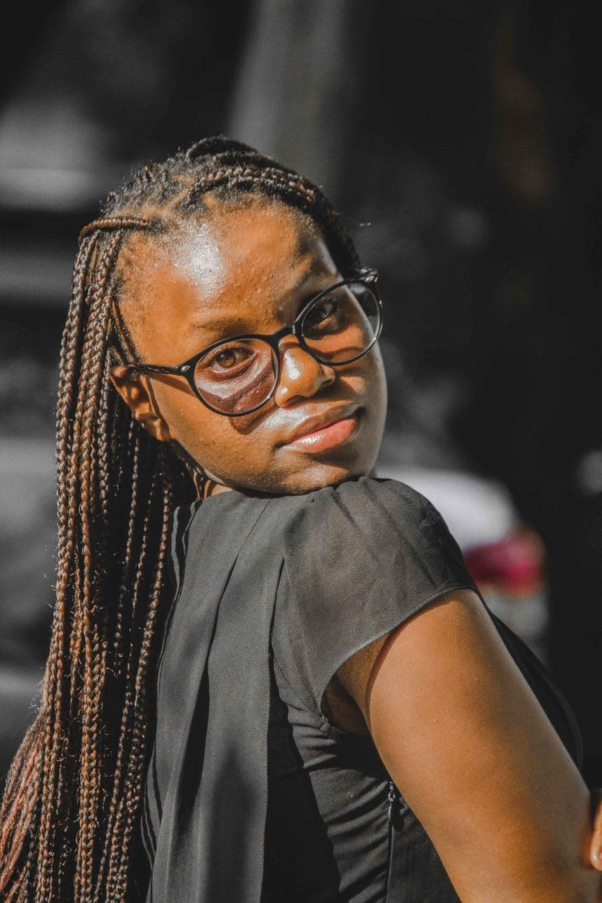 Close-up portrait of a young woman with eyeglasses and knotless braids, exuding confidence outdoors.