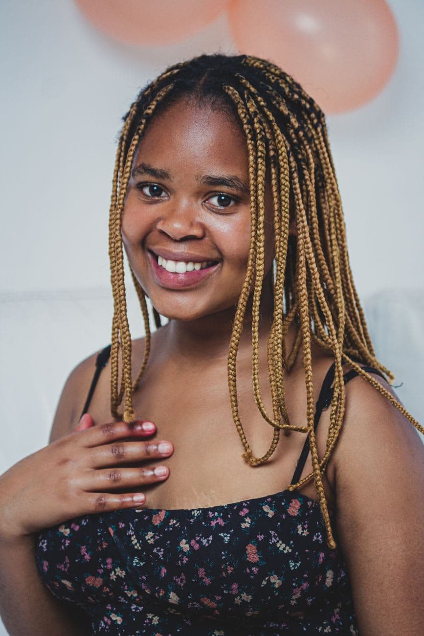 Portrait of a smiling woman with braided hair, wearing a floral top, hand on chest.