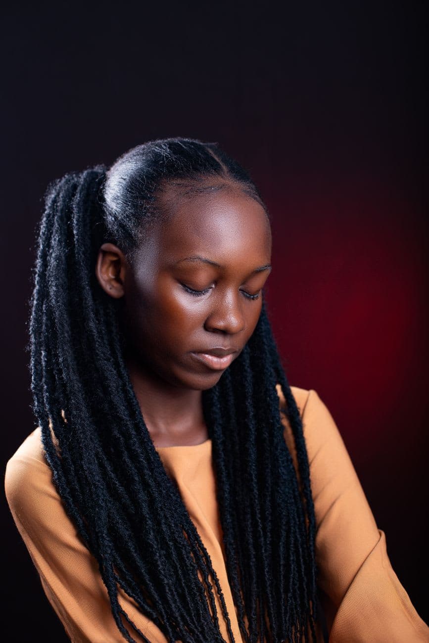 Elegant studio portrait of a woman with dreadlocks wearing a mustard dress against a dark background.