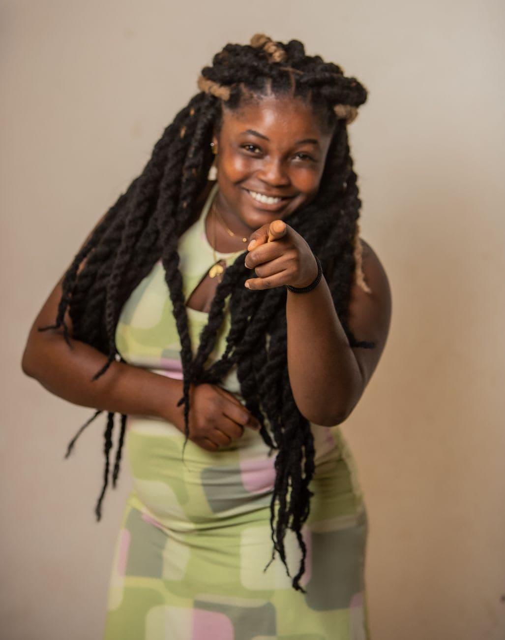 A joyful woman with long locs and a colorful dress points forward in a cheerful pose.