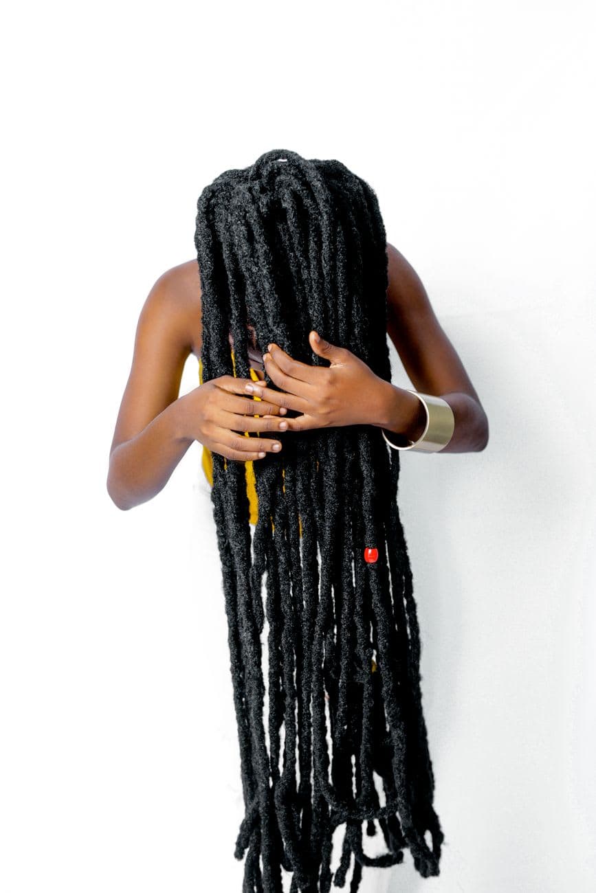 A woman with long black dreadlocks poses artistically against a white backdrop, showcasing unique hairstyle.