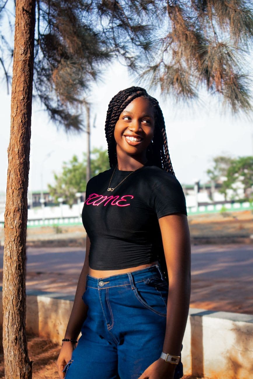 A cheerful young woman with braided hair smiling in a sunny outdoor setting.