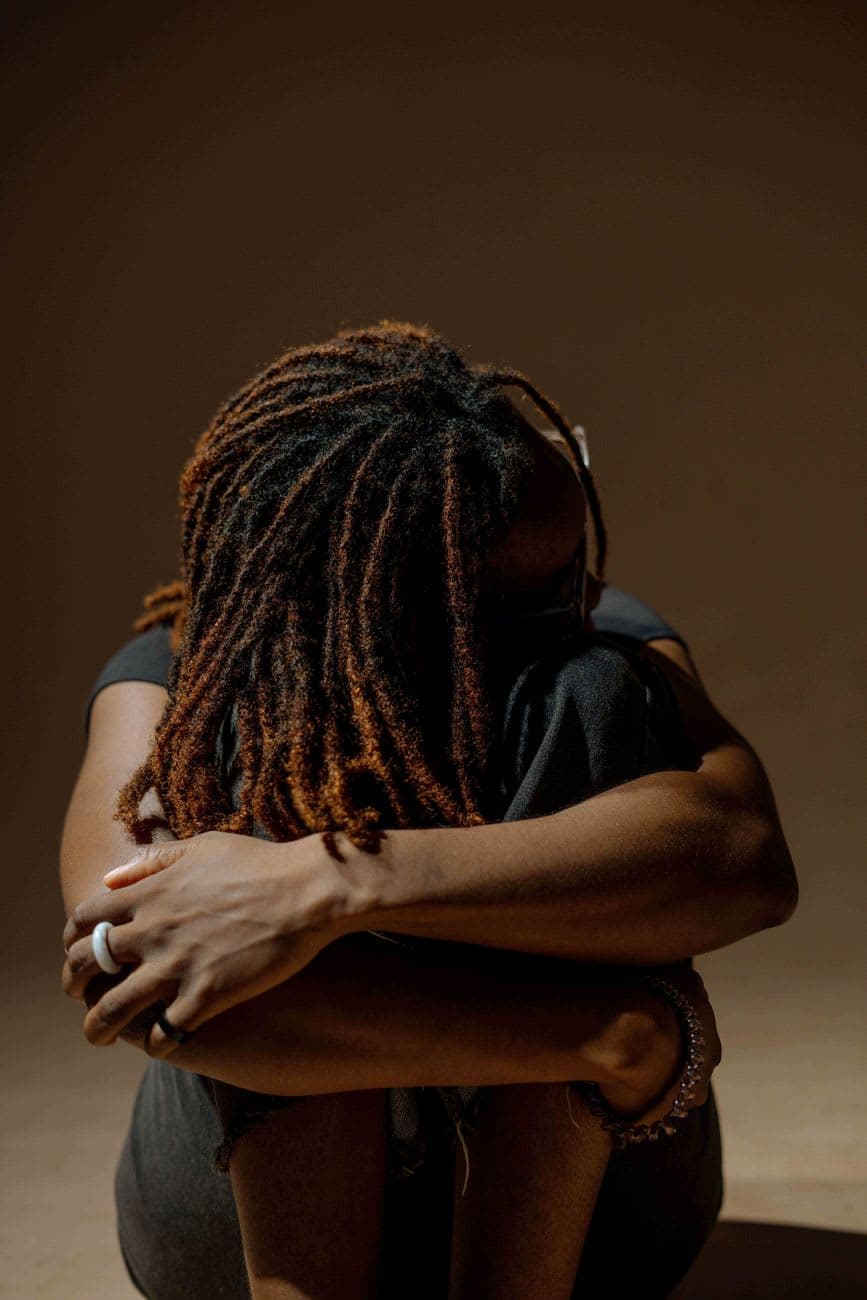 A woman with dreadlocks embraces herself in a thoughtful pose. Captured with warm tones, this portrait conveys introspection.