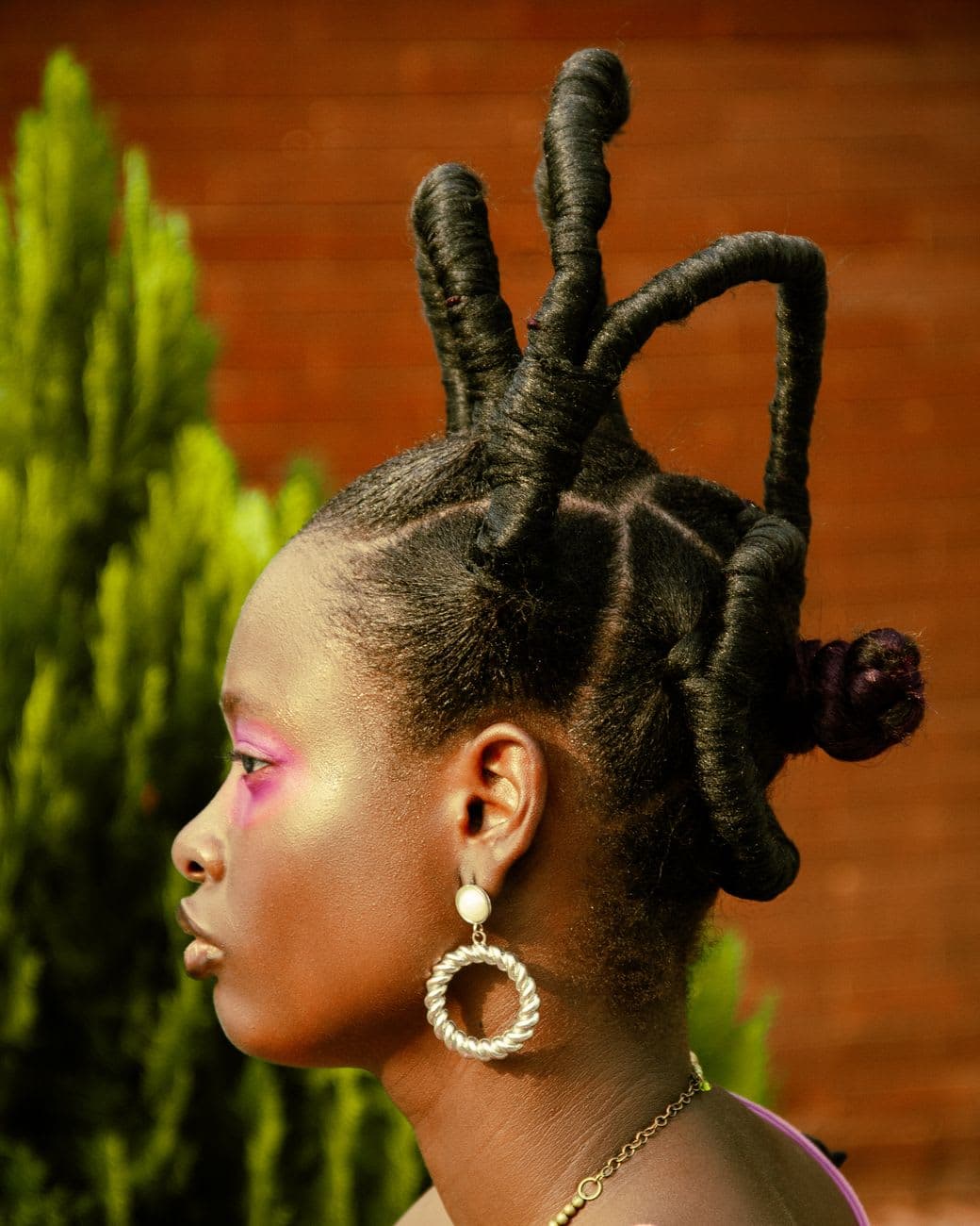 A woman with a creative twisted hairstyle and bold makeup standing outdoors. Vivid colors enhance her striking look.