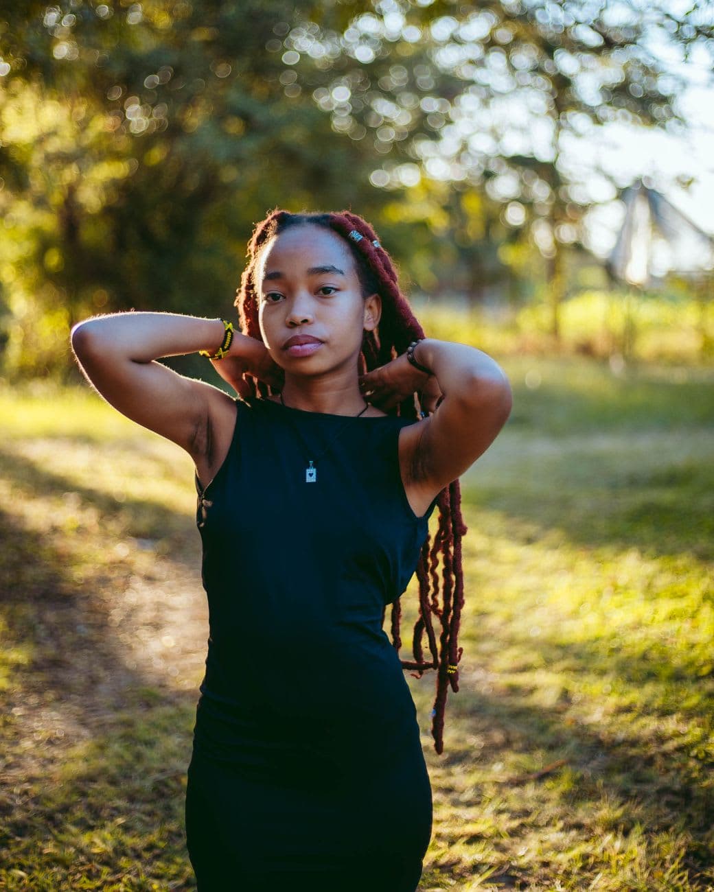 A fashionable young woman with long braids poses confidently in an outdoor setting during sunset.