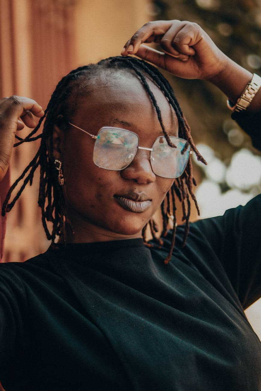 Portrait of a fashionable woman with dreadlocks and glasses posing outdoors in sunlight.