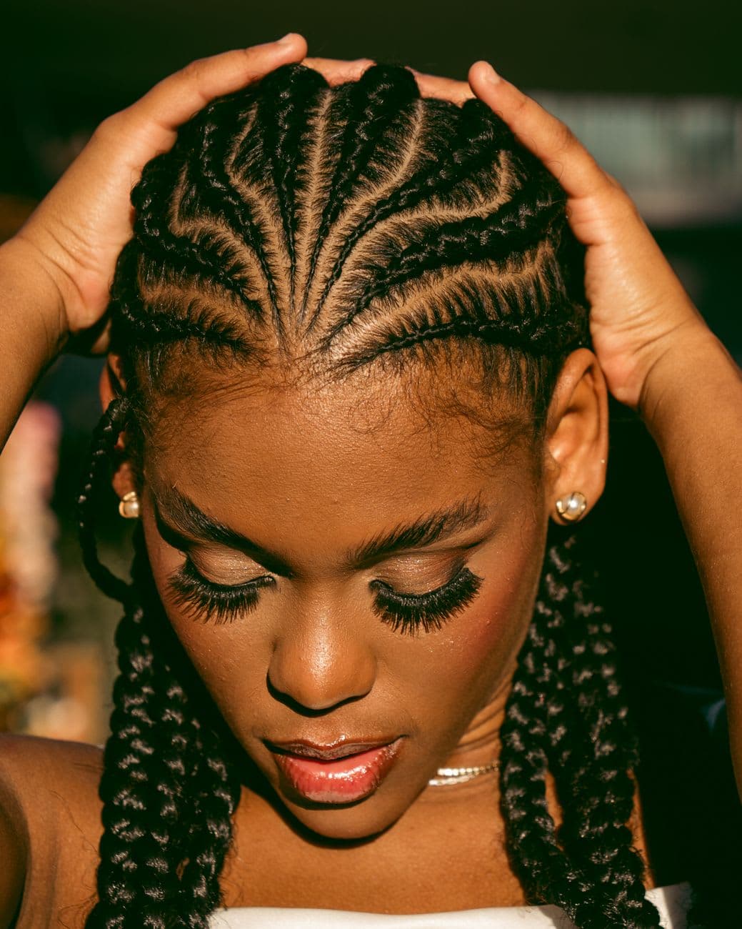 A thoughtful close-up portrait of a woman wearing cornrows with a warm, natural light backdrop.