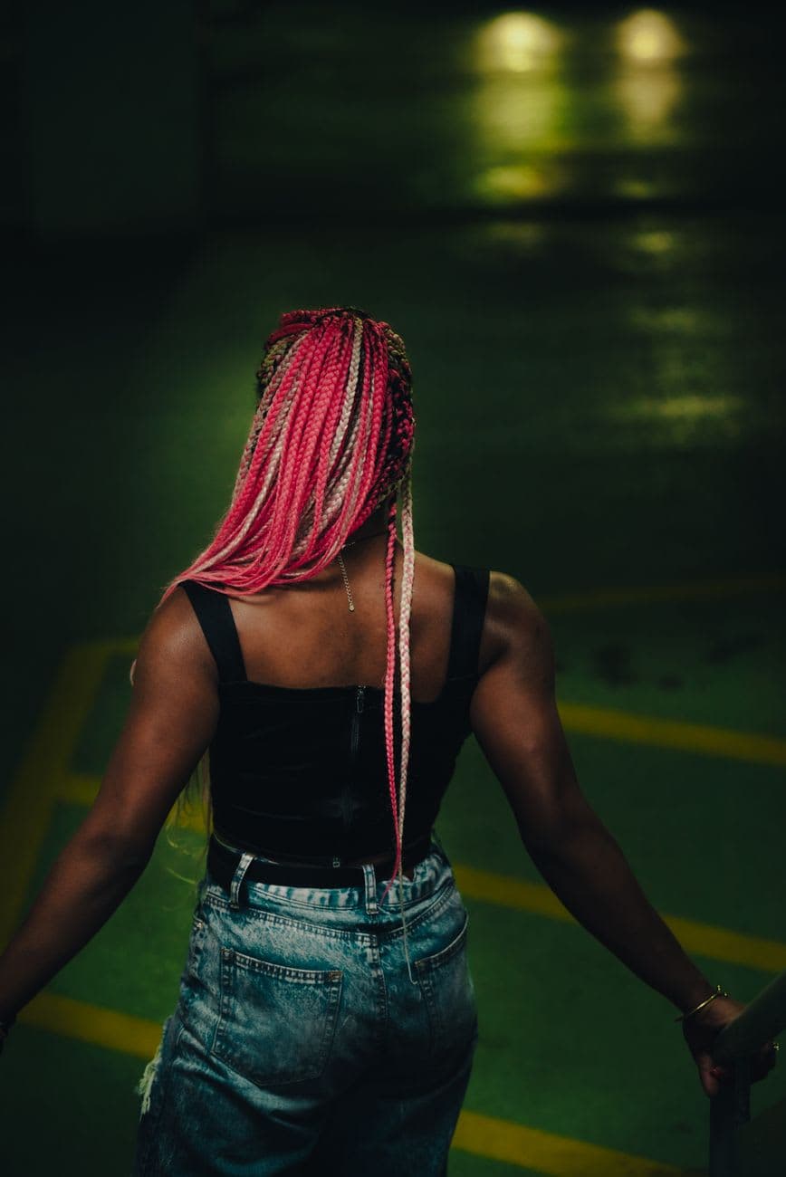 Stylish woman with pink braids in a tank top stands indoors, showcasing vibrant fashion.