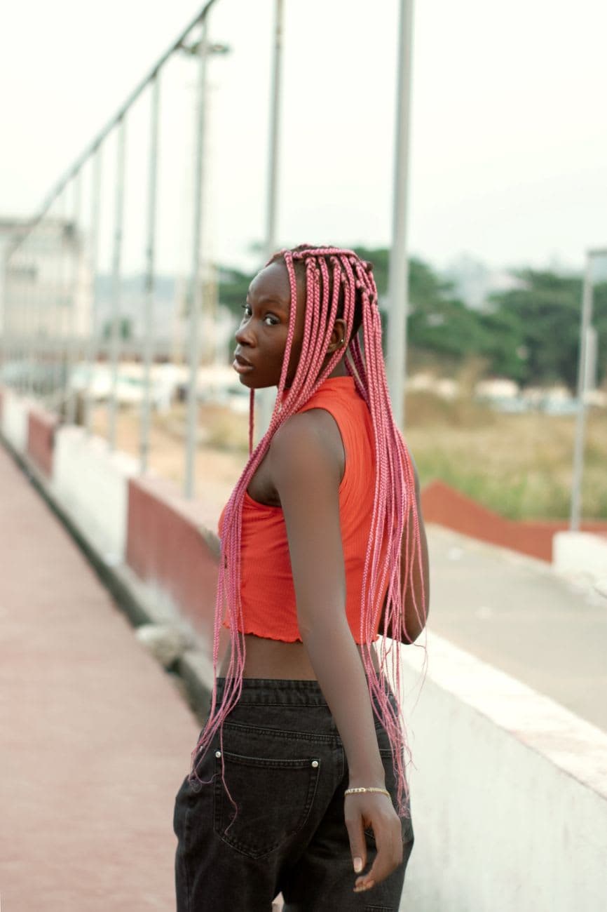 A fashionable woman with long pink braided hair looks back while walking outdoors in a vibrant red crop top.