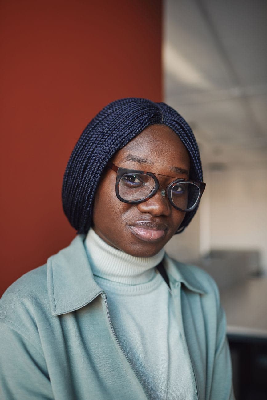 Close-up portrait of a woman with glasses and braided hair in a stylish outfit indoors.