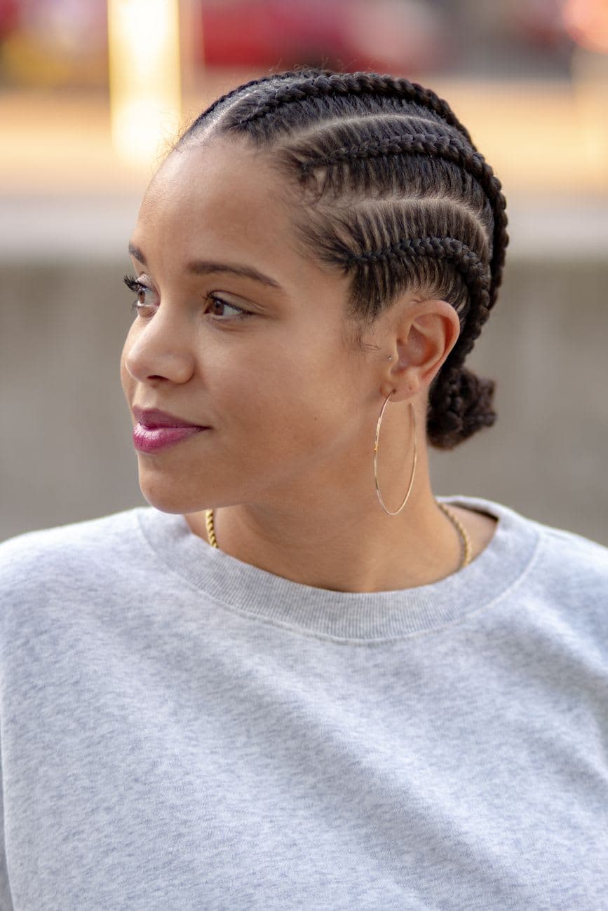 A woman with braided hair and loop earrings gazes afar, wearing a gray sweater outdoors.